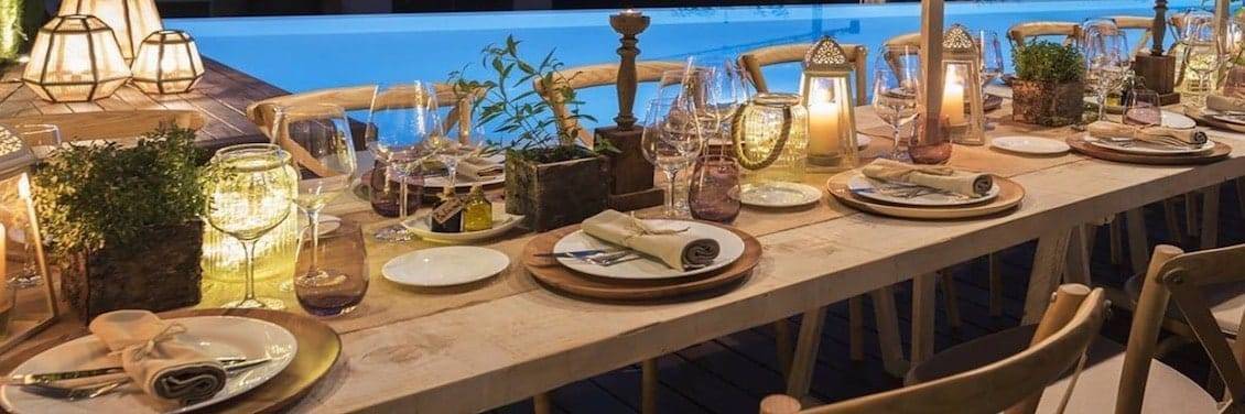 Mesa rústica al aire libre con iluminación cálida, decorada con faroles, copas de cristal, vajilla en tonos neutros y detalles naturales junto a una piscina.